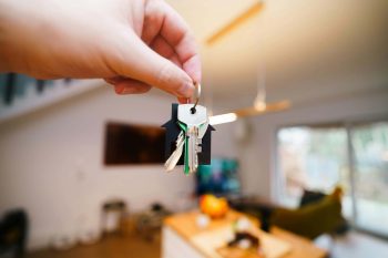 Hand holding rental property keys with a home keychain in front of a furnished living room — representing the flooring decisions Texas landlords face when choosing the most durable carpet for tenant turnover